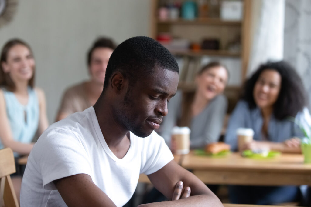 A photo depicting a dejected Black man sitting at a table by himself while a group of people laugh in the background.