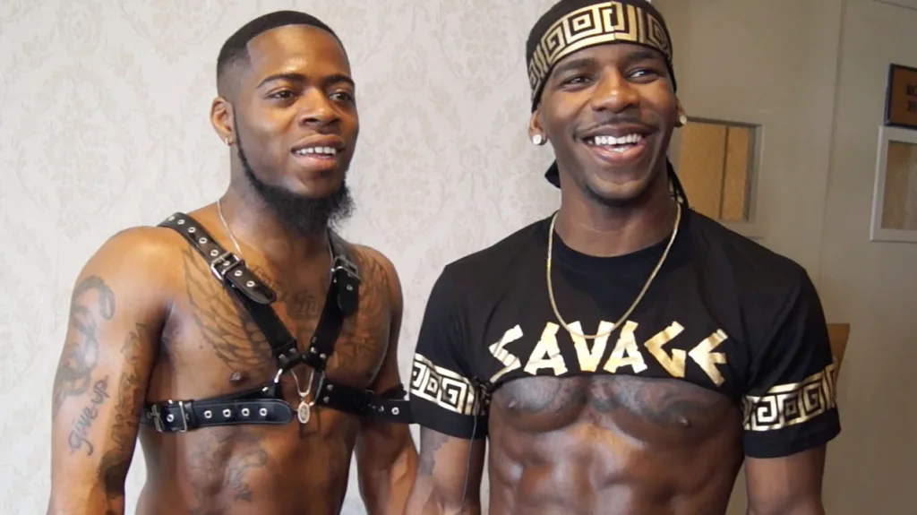 Two smiling Black men, one wearing a harness, the other wearing a cropped shirt that says "savage"