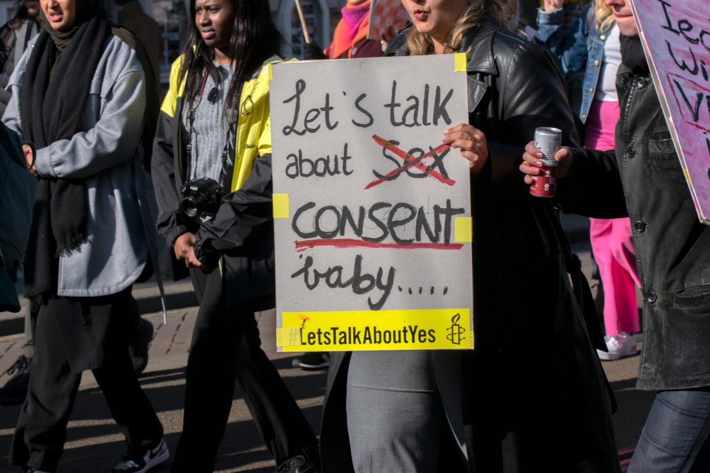 Woman holding sign that says "Let's talk about sex consent baby..." with "sex" crossed out indicating an emphasis on consent-seeking behavior