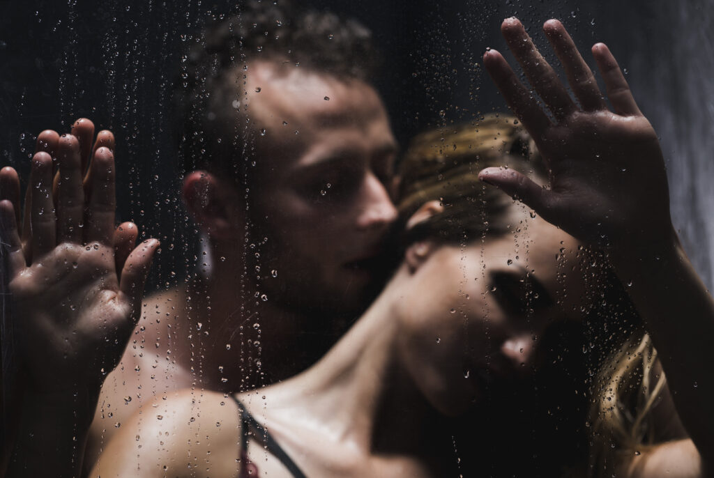 Man standing behind woman talking in her ear as he pins her hands to a glass door.