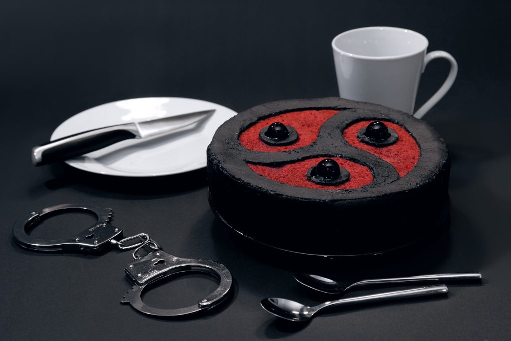 Cake in the shape of a triskelion, surrounded by a mug, two spoons, handcuffs, a plate and a knife.