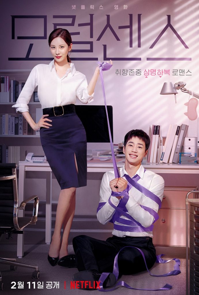 Image of a seated, smiling Korean man in business clothes wrapped in bondage tape with a Korean woman in business clothes stands beside him holding part of the tape.